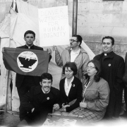 Hispanic and white men and women rally for improved living conditions for migrant workers at the Capitol Building in Denver, Colorado. A priest holds a flag that depicts an eagle and reads: "Huelga U. F. W. O. C. AFL-CIO Delano." Another man holds a sign that reads: "Denver Witnesses for Human Dignity."