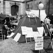 Migrant workers and other protestors camp near possibly the Colorado State Capitol building in Denver, Colorado. They sit near a group of tents. Handwritten signs read: "We Welcome Martin Luther King Sr.," "Boycott Scab Grapes," and "Support Farm Labor Legislation."