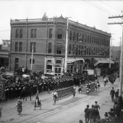 Chinese men march in a parade, part of the Festival of Mountain and Plain, on 16th (Sixteenth) Street in downtown Denver, Colorado. They carry elaborately decorated banners. Spectators line the street. The Waugh Building is nearby.
