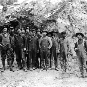 Miners, some possibly Native American or Asian, pose outdoors in Ouray County, Colorado. Some men hold lunch boxes.
