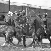 Cast members of Buffalo Bill's Wild West Show duel with swords while mounted on horses. They wear fencing outfits, mesh masks and helmets.