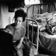 A nun seated on the edge of a bed listens to an unidentified migrant worker. A man and a young boy, lying in a bed behind her, look on.