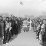Native American (Ute) and white men and women stand in rows opposite each other probably for the traditional Bear Dance, probably in western Colorado. Shows A U.S. flag and possibly a school in the distance.