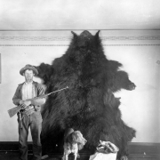 A man holds a rifle and stands next to a grizzly pelt that hangs from a wall in a residence in Arizona. A dog stands in front of the pelt; the grizzly skull sits on a box next to the dog.