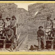 View of a Chinese construction crew on the Union Pacific Railroad, the men sit and stand next to a white man on a railroad handcar near a rock formation.