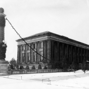 View of the Denver Public Library at Colfax Avenue and Bannock Street in the Civic Center neighborhood of Denver, Colorado. The Greek Revival style building has columns and is decorated with greenery for Christmas. A column is in Civic Center Park nearby. Snow covers the ground.