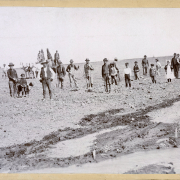 Farm workers, men and boys, pose with two well dressed men, a boy and a girl, in a plowed farm field in probably western Colorado. The farm workers wear work clothes and hats, and hold shovels and tools. The boys wear knickers and caps; one boy stands beside a dog. The girl wears a pinafore and a bonnet. An irrigation ditch and wet soil are near the people. Two men pose with trees, probably transported for planting, and a barrel on a wagon pulled by horses.