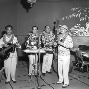 Interior photograph of four musicians playing music (possibly) at the Lotus Lounge for the Chinese New Year Celebration. Each of the musicians wears a floral shirt; they play the guitar, keyboard, cello, and the ukulele.