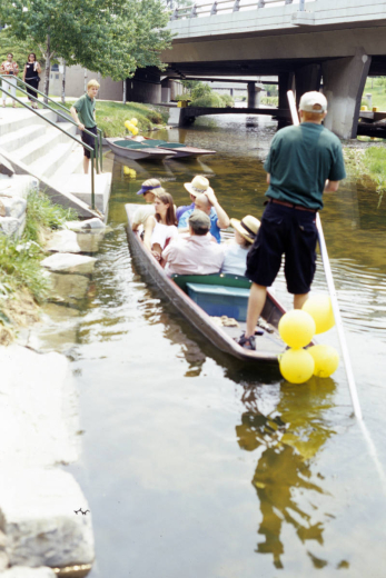 View of punting boats from the company Venice on the Creek near 14th and Larimer in the Larimer Square Historic District in the Union Station neighborhood, Denver, Colorado. A man in a green shirt and black shorts stands in the boat and punts with a white pole. Six passengers sit in the punt, which is decorated with four yellow balloons. Two empty punts are tied up nearby, and a man is on stairs that lead to the creek.
