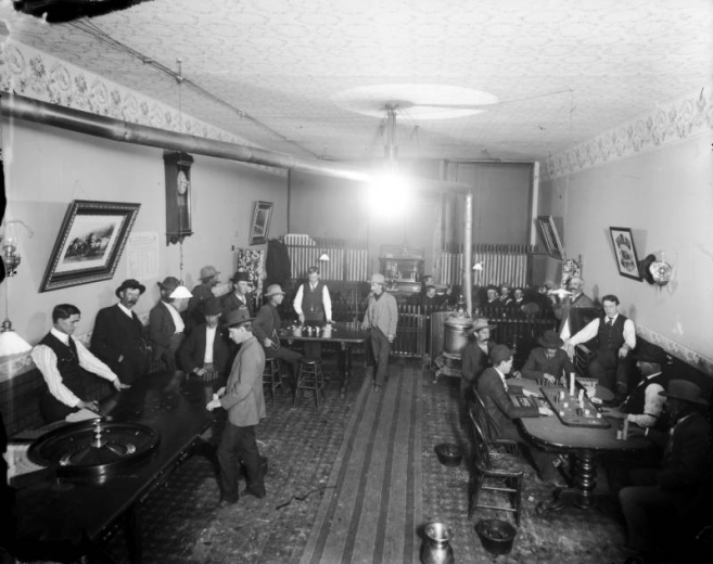 Interior view of a saloon in Leadville (Lake County), Colorado; shows men gambling, a roulette wheel, gaming tables, playing cards, poker chips, croupiers, tables with playing cards and poker dealers, and a tray of glasses. Decor includes floral wallpaper, a wainscot, framed pictures and a clock. A cast iron stove is by spittoons; pool cues are in back.
