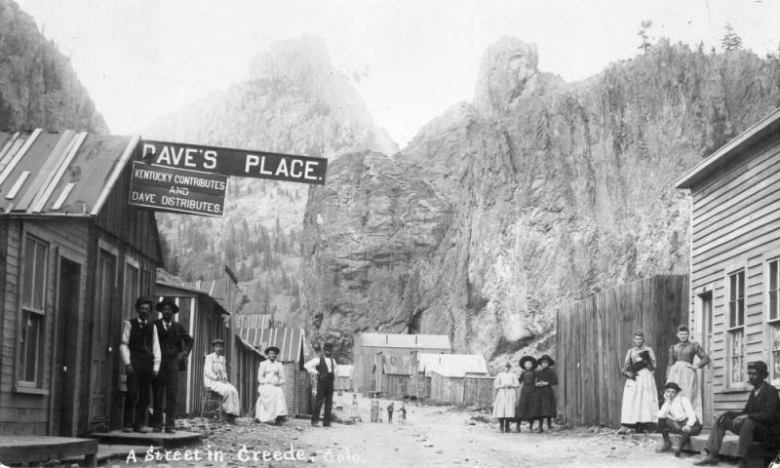 Men, women, and children stand or sit and pose on a street in Creede (Mineral County), Colorado. White and Black (African American) men wear suits or vests and hats. Women wear dresses and aprons; a woman holds a dog. A sign on a business reads: "Dave's Place, Kentucky Contributes and Dave Distributes." Commercial buildings have false fronts and gabled roofs with tar paper.