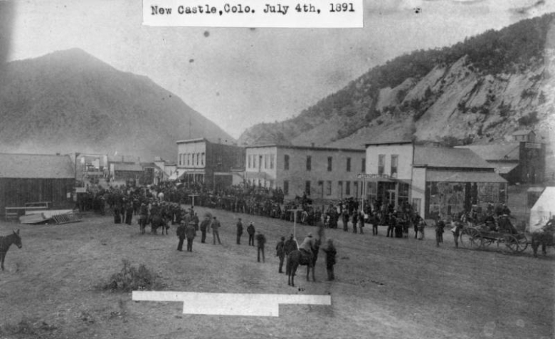 Spectators crowd the edges of Main Street on July 4th in New Castle, Garfield County, Colorado. Horses, wagons and people are in the dirt thoroughfare, and two-story commercial buildings are behind them. A mining shed is against the rocky slope in the background; signs read: "Colorado," "Furniture," and "Saloon."