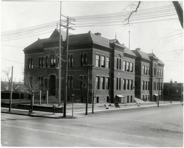 Photograph of one of the early locations for the Emily Griffith Opportunity School located in Denver, Colorado.  Two young boys sit on the curb outside of the school.