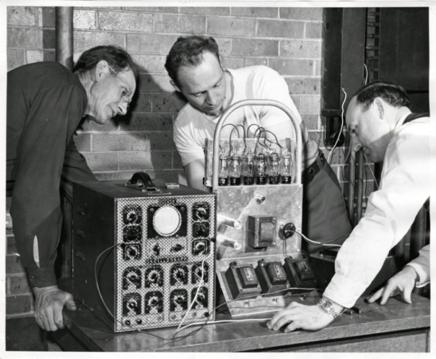 Photograph of three men during a radio repair class at the Emily Griffith Opportunity School located in Denver, Colorado.  Two men watch while a man in the center stands behind the machine working on it.