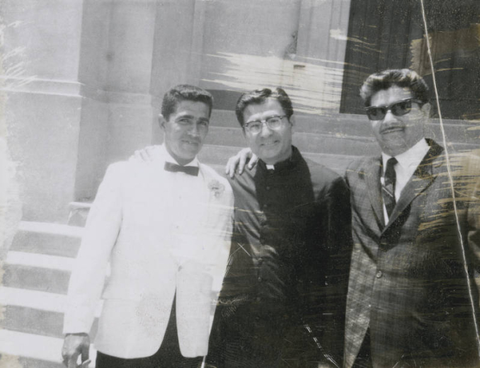 Jim Herrera (Skip), Father Pete Garcia and Bobby Baca (Nimbo) stand near the steps of St. Cajetan's church in the Auraria neighborhood, Denver, Colorado. Jim Herrera wears a white jacket and bow tie. Father Garcia wears eyeglasses, a cassock and clerical collar, and Bobby Baca wears a plaid jacket, tie and dark glasses.