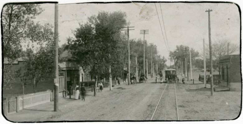 Photograph of a street in the Globeville neighborhood of Denver, Colorado.  Pictured is an electric street car in the street, with various individuals walking along the sidewalks. Date estimated based on start of electric trolley service in this area (1908) and street scene.