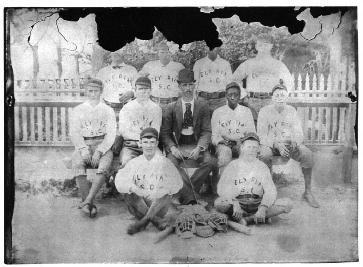 Photograph of a Elyria's baseball team.  The team sits in three rows, two on the ground, five on a bench, and four standing behind.  All wear their uniforms.  The upper portion of the photograph has damage and some of the faces can not been seen