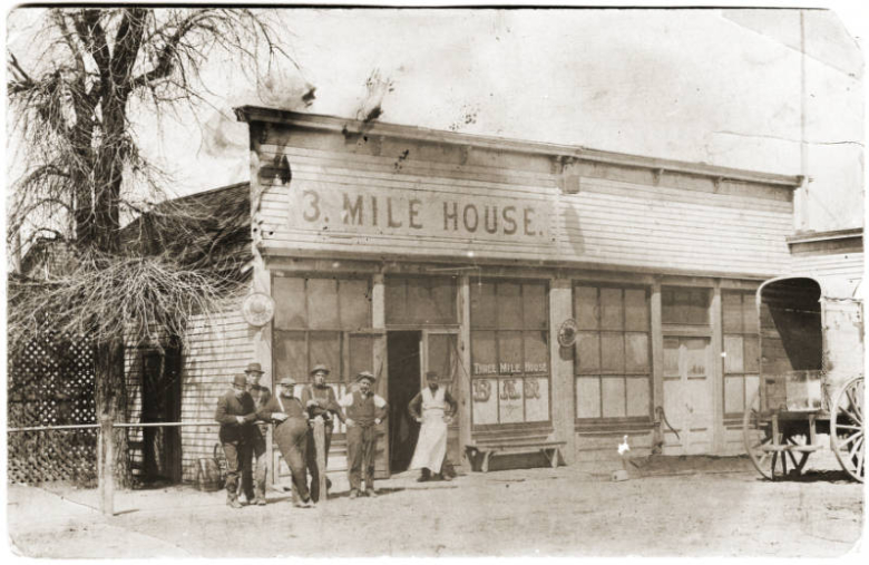 Photograph found on a postcard of the Three Mile House located in the Elyria-Swansea neighborhood of Denver, Colorado.  Several men lean up against a hitching rail on the left side of the door.  A man in a long white apron stands in front of the door.  A wagon is seen on the right hand side of the photograph.  Built before 1881, Three Mile House was originally owned by Philip Kiefaber and his wife Elizabeth.  Three Mile House was comprised of a farm, dairy, inn, saloon and social hall.