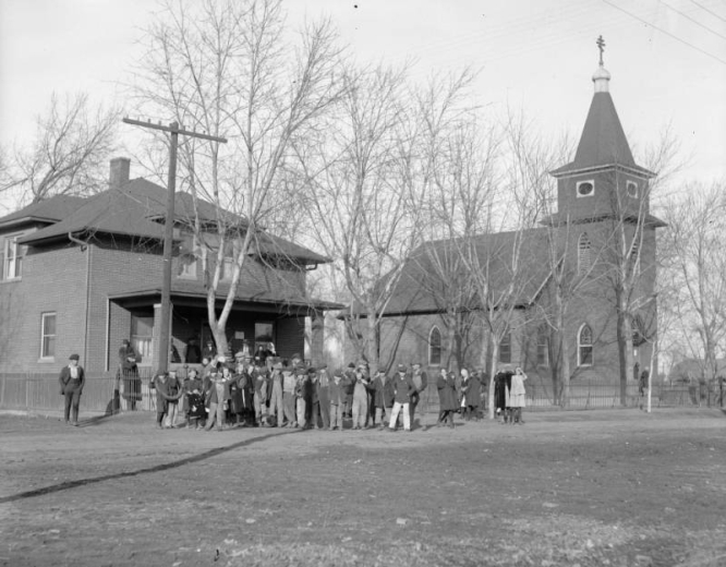 A group of people pose near a two-story, brick house and the Holy Transfiguration Russian Orthodox Church, located at 47th (Forty Seventh) and Logan Streets in the Globeville neighborhood of Denver, Colorado. The church has a gabled roof, bell tower with a cross and arched, painted windows. Shows trees and a fence.