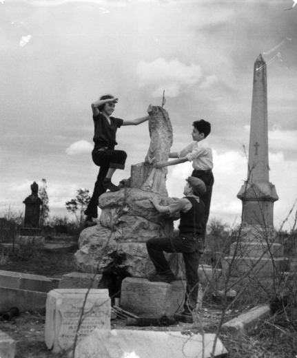 Three children, two boys and a girl, play on a stone momument among the tombstones and monuments at Mount Calvary Cemetery, the Catholic section of Mount Prospect Cemetery (later Denver Botanic Gardens), in the Capitol Hill neighborhood of the City and County of Denver, Colorado.  The girl stands near the top of the monument and holds her hand above her eyes to view the surrounds.