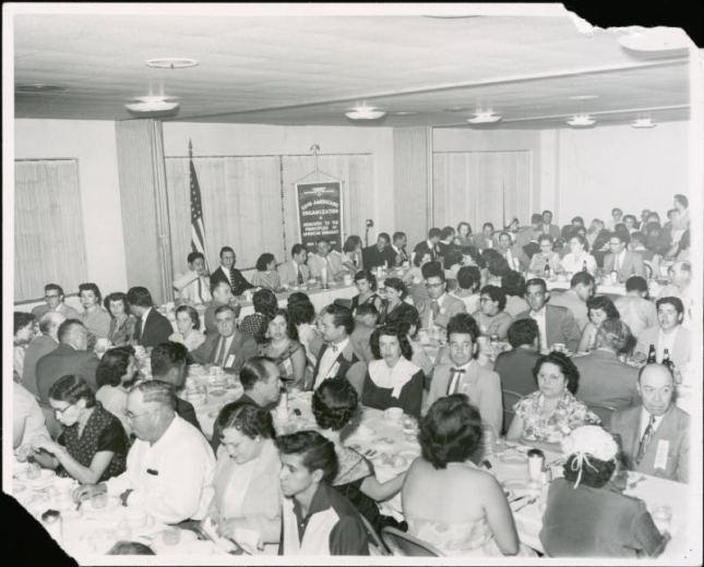 View of a Good Americans Organization (G.A.O.) banquet probably in Denver, Colorado. Well dressed men and women sit at banquet tables. There is a head table with men and women and a priest at the front of the room. An American flag is beside the head table and a banner hangs on the wall. Lettering on the banner reads: "Good Americans Organization, Dedicated to the Principles of American Democracy." "Org. ? 1954." An inverted triangle on the banner has the letters: "CO GAO."
