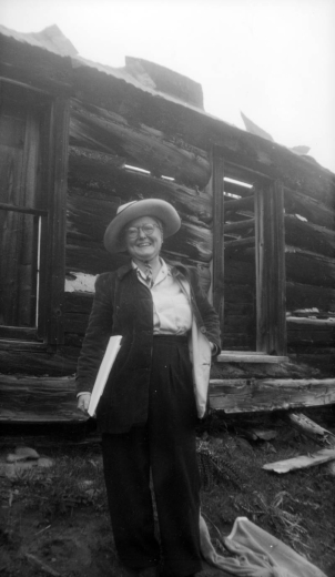 Muriel S. Wolle poses by a log cabin near Boulder, Boulder County, Colorado, wearing pants, a button-down shirt, an overcoat, glasses, and a cowboy hat. She carries a sketch pad.