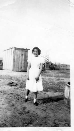 Magdalena Gallegos stands in a corral in front of an outhouse, at her great-aunt Romanita's ranch, in Keenesburg (Weld County), Colorado. She was 13 or 14 at the time. She wears a white dress with puffed sleeves and loafers and white socks. A wooden barrel and a loading chute are nearby.