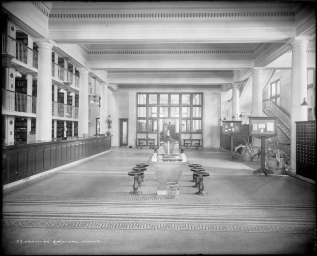 Interior view of (Carnegie) Denver Public Library, Denver, Colorado; shows lobby with circulation desk, bookshelves on three floors, card catalogs, center wooden tables with metal stools, geometric pattern bands in tile floor, agave plants in barrels and informational sign boards.