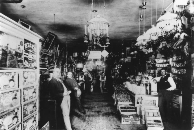 The Sauer McShane Mercantile Company on Eureka Street is crowded with merchandise, including boxes of soap, baking powder, fruits, nuts, and chandeliers. The shopkeeper, in a vest, is behind the counter. Men lean against the sales counters looking at the camera. A "Burnette's Extracts" sign hangs from the chandelier in the middle of the room. A box printed with "Pride of Soap Denver" is on the floor.