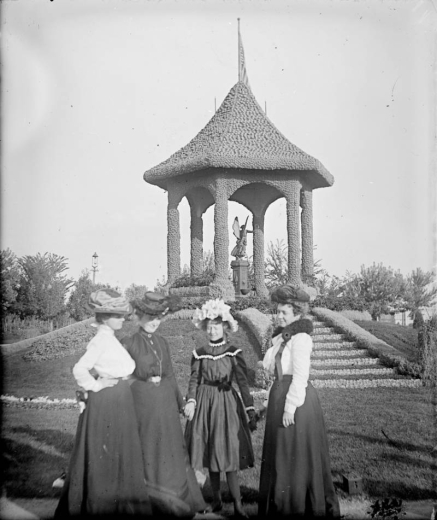 A group of women stand near the Spanish-American Memorial in City Park, Denver, Colorado. They hold hands and wear hats decorated with flowers. An angel statue is in the gazebo.