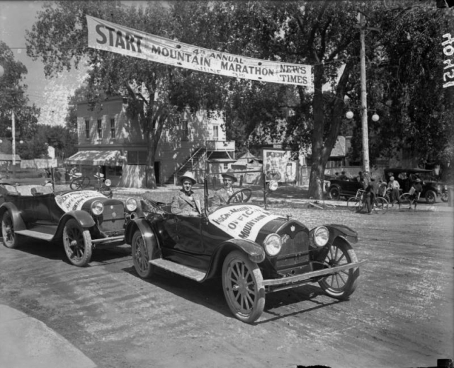The start of the 4th Annual Mountain Marathon sponsored by the Rocky Mountain News and the Denver Times, Denver, Colorado. A banner that hangs across the street reads: "Start. 4th Annual Mountain Marathon June 3rd. News-Times." Shows bystanders, a bicyclist and two automobiles, both with banners across the hoods. Two men pose in a Briscoe automobile.