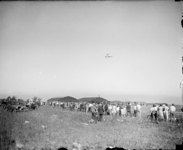 People and cars crowd Lowry Field in Denver, Colorado, watching Charles Lindbergh's Spirit of Saint Louis in flight. A boy has a bicycle.