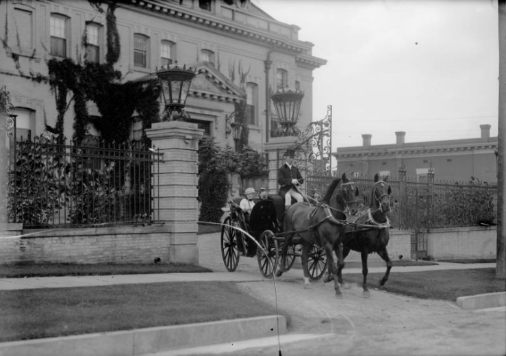 View shows Mrs. Crawford Hill riding in a carriage being driven by a chauffeur, and pulled by a two-horse team, Denver, Colorado. Her residence is behind her.