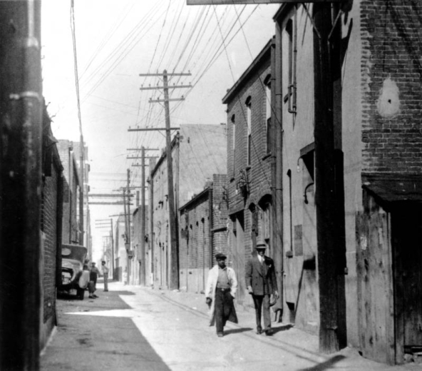 Men walk on Hop Alley (between Wazee and Blake Streets) in Denver, Colorado. Another group of men sit near an automobile.