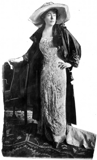 Studio portrait of Margaret "Molly" Tobin Brown. She wears a dress decorated with fringe, a coat with fur collar, and a wide-brimmed hat.