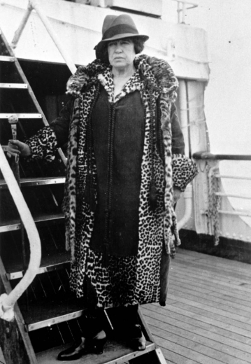 Margaret "Molly" Tobin Brown poses on the steps of a ship. She wears a leopard trimmed coat, dress, a wide-brimmed hat, and holds a swagger stick.