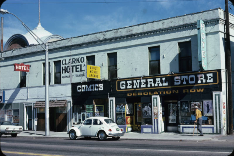 View of the Clarko Hotel at 813 East Colfax in the North Capitol Hill neighborhood of Denver, Colorado. This is a two-story twentieth-century brick building that has been painted white. The entrances to the businesses are recessed. There is a cornice with a row of dentils and a parapet.