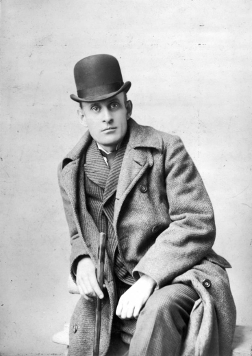 Studio portrait of Eugene Field in San Francisco, California. He holds a cane and wears an overcoat and bowler hat.