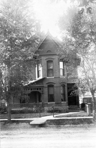 View of a two story house designed and built by Orlando Scoby on 2743 Curtis Street in the Curtis Park Historic Districts in Denver, Colorado. The brick house has a porch with decorative wood trim, a pitched roof, and iron fence. A flagstone carriage stoop is in front of the house.