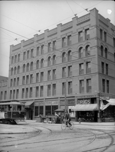 View of the Oxford Hotel and corner Drug Store on 17th (Seventeenth) Street in Denver, Colorado. Business signs read: "Drugs, Cigars, Candies" "Austin's Candies", and "La Matilde." Shows: Denver Tramway trolley 17th Ave. #48, wagon, man on bicycle.
