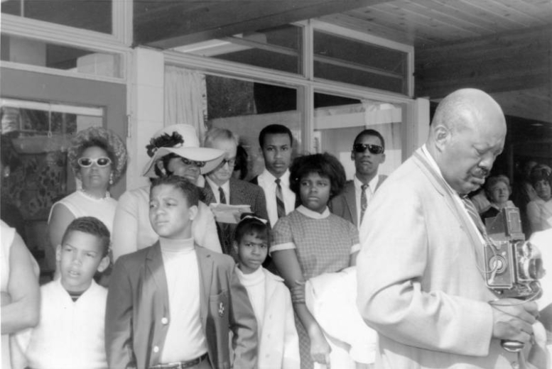 Men, women, and children stand near a building during the George Washington Carver Day Nursery dedication at 2260 Humboldt Street in the City Park West neighborhood of Denver, Colorado. Burnis McCloud stands by a camera near the group.