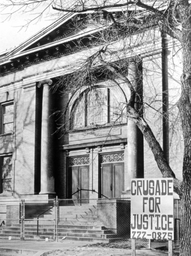 A front view of the Crusade for Justice building located at 1567 Downing Street in Denver, Colorado. The building was originally the Calvary Baptist Church. It features columns with scrolls on top, a semi-circular window above a set of double doors and steps. A wire fences blocks the entrance way; a billboard sign that reads: "Crusade for Justice 222-0825" is in front of the fence.