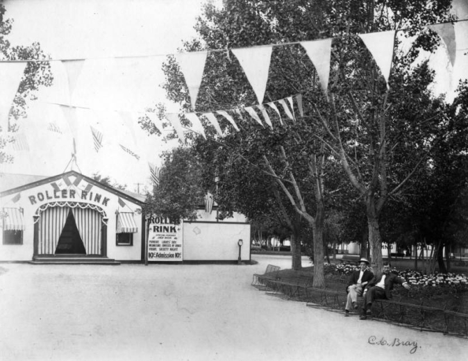 View of Manhattan Beach amusement park in Denver, Colorado; shows men on a bench by landscaping, and a building with signs: "Roller Rink Special Features Each Week, Monday, Ladie's Day, Wednesday, Contests of Grace, Friday, Society Night, 10 Cents Admission." A scale is to the side; United States flags hang from ropes.