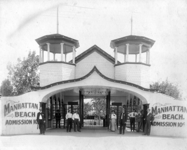 View of the Manhattan Beach amusement park entrance in Denver, Colorado; shows people standing by an arched gate with towers, shingle imbrication, and lettering; "Manhattan Beach Admission 10 cents." A man wears a police uniform with helmet; a woman holds a small handbag.