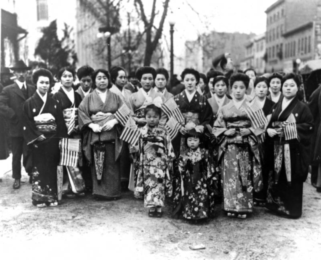 A group of Japanese-American women and young girls dressed in kimonos pose in the middle of a street, possibly in Denver, Colorado. Some of the women hold miniature American flags. A man stands next to the group; he wears a suit, tie and hat. Front row (behind young girls), first from left is Mrs. Reverend Ono; second from left is Mrs. N. Hokasono. Front row third from right is Mrs. Y. Hayano and second row, first from right is Mrs. M. Otsuki.