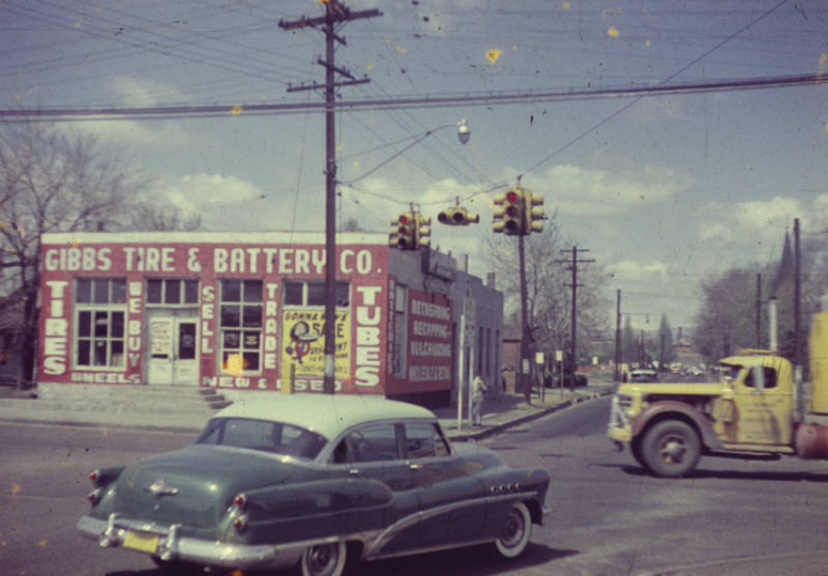 View of Gibbs Tire and Battery Company at 4589 Washington Street in the Globeville neighborhood of Denver, Colorado. An automobile and a truck make their way through the intersection of Washington Street and 46th Avenue. Signs on the building read: "Tires We Buy Sell Trade Tubes."