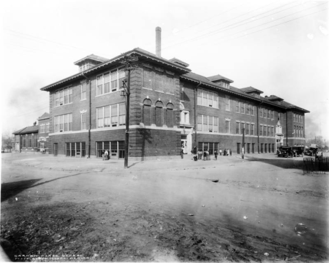 View of Garden Place School, 44th (forty-fourth) Avenue and Lincoln Streets, Denver, Colorado; shows a brick building with quoins, projecting eaves, exposed rafters, and dormers. Boys and girls play, cars are parked.