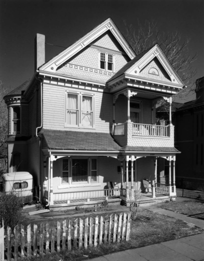 View of 2825 Curtis Street in the Curtis Park neighborhood (Five Points) in Denver, Colorado; shows a frame house with shingle imbrication, a covered porch and balcony, and round bay windows. A camp trailer is to the side.