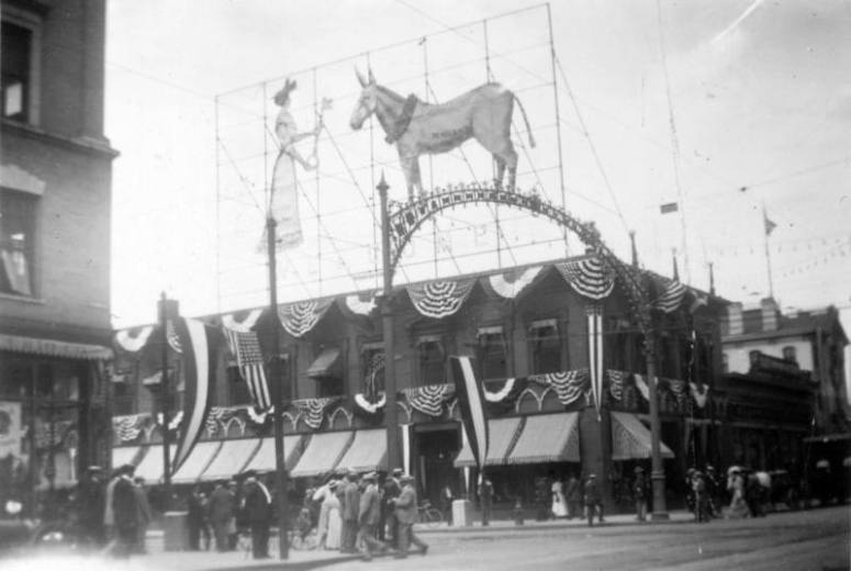 Pedestrians stand near the Aspen Building (while it was home to the Gas and Electric Company) on the corner of 17th (Seventeenth) Street and Tremont Place in downtown Denver, Colorado. The building is decorated with bunting and has a lighted sign of a woman who holds a key out to a donkey on the roof. Lettering on the donkey reads: "Democracy." The electric sign is near a decorative metal arch that spans the street, both were erected for the Democratic National Convention.