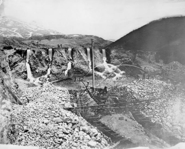 View of a hydraulic gold mine in Breckenridge, Summit County, Colorado; shows eroded embankments, falling water, jets of water from "monitor" nozzles, sluices, flumes, a crane, and miners. Mountains are in the background.